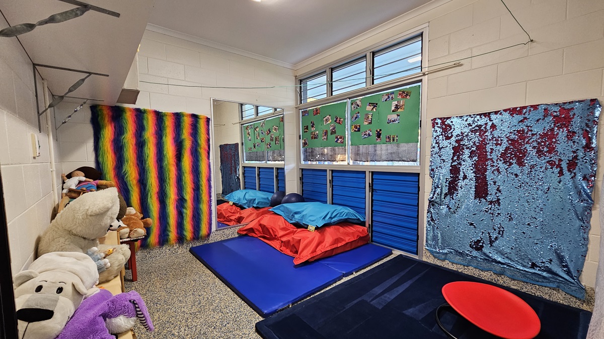 On the left, there are shelves filled with stuffed animals and toys, next to a wall covered with a rainbow-striped fabric. On the right, there is a wall with a blue and silver sequined fabric. In the middle, there are blue mats on the floor with red and blue cushions, and windows above with green boards displaying photos. The floor is speckled grey carpet, and the ceiling is white with recessed lighting.