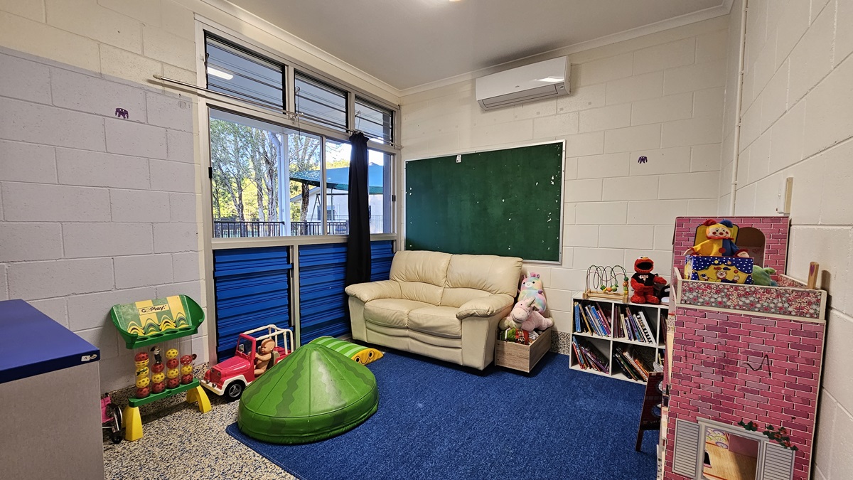 On the left, there is a blue and white wall with windows above, a green toy shelf with books, a pink toy truck, and a green play structure. On the right, there is a white wall with a pink dollhouse, a bookshelf with toys, and a green chalkboard. In the middle, there is a beige sofa with stuffed animals on it. The floor is covered with blue carpet and speckled grey tiles. The ceiling is white with recessed lighting and an air conditioning unit.