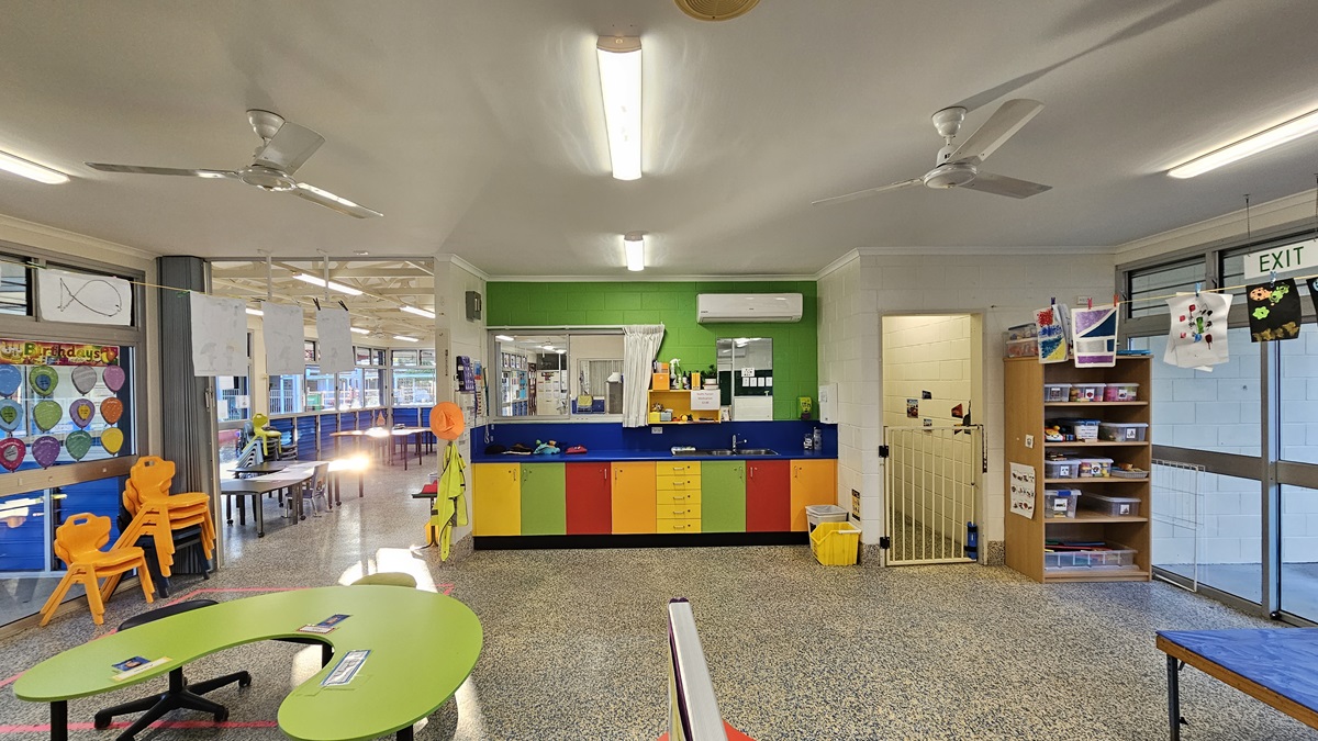 On the left, there are colourful chairs around a green table, a wall with circular artwork, and a doorway leading to another room with tables and chairs. On the right, there is a bookshelf with toys and books, a door with an "EXIT" sign, and children's artwork hanging above. In the middle, there is a counter with colourful cabinets below and a green wall above, featuring a sink and an air conditioning. The floor is speckled grey, and the ceiling is white with fluorescent lights and ceiling fans.