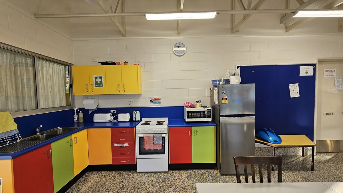 On the left, there is a kitchen area with a sink, a drying rack, and colourful cabinets in yellow, red, and green. On the right, there is a stainless steel refrigerator, a microwave on a blue counter, and a small table with a blue helmet on it. In the middle, there is a white stove with an oven, and a clock on the wall above. The floor is speckled grey, and the ceiling is white with exposed beams and fluorescent lights.