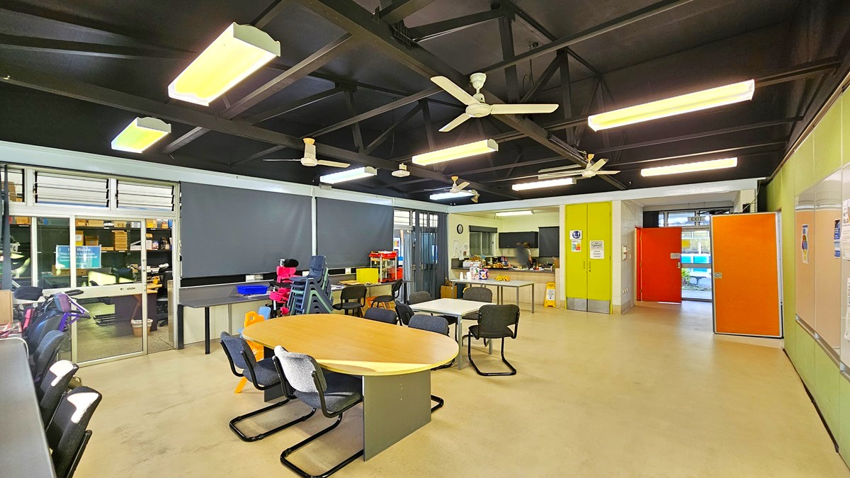 On the left, there are windows with blinds, a black desk with a computer, and a stack of colourful chairs. On the right, there is a green wall with a bulletin board and an orange door. In the middle, there is a round wooden table with black chairs around it. The background features a kitchen area with a sink, cabinets, and a doorway. The floor is light-coloured concrete, and the ceiling is black with exposed beams, fluorescent lights, and ceiling fans.