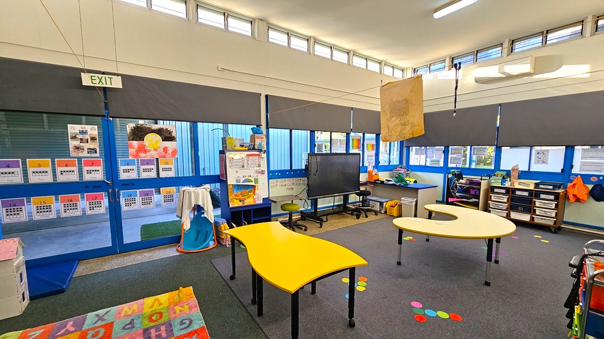 On the left, there are blue-framed windows with black blinds, an "EXIT" sign above a door, and colourful posters on the wall. On the right, there are more blue-framed windows with black blinds, a whiteboard, and a set of cubbies with various items. In the middle, there are two yellow tables with curved edges, a black television on a stand, and colourful floor markers. The floor is a mix of grey carpet and a colourful alphabet rug. The ceiling is white with recessed lighting and exposed beams.