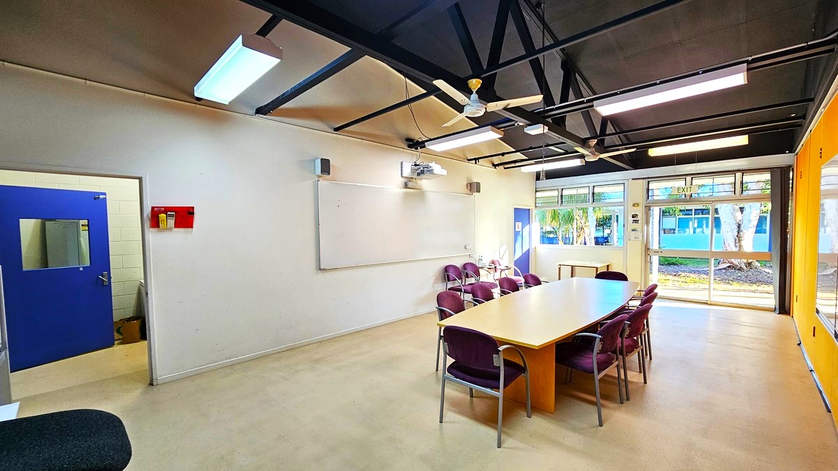 On the left, there is a white wall with a blue door featuring a small window, a red fire alarm box, and a whiteboard with speakers above it. On the right, there are large windows with a view of trees and a building outside, and an orange wall with a blue door. In the middle, there is a long wooden table surrounded by purple chairs. The floor is light-coloured concrete, and the ceiling is black with exposed beams, fluorescent lights, and ceiling fans.