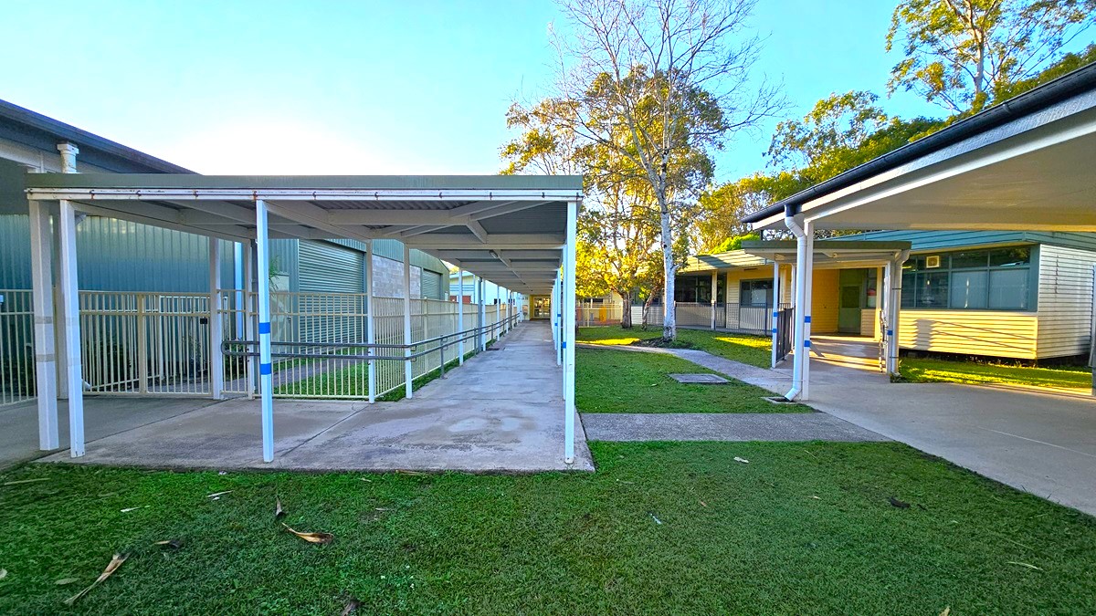 On the left, there is a covered walkway with white railings and a green building with vertical panels. On the right, there is another covered walkway leading to a building with large windows and a yellow wall. In the middle, there is a concrete path flanked by grass, leading to a tree and more buildings in the background. The floor is concrete, and the ceiling of the walkways is white with metal supports.