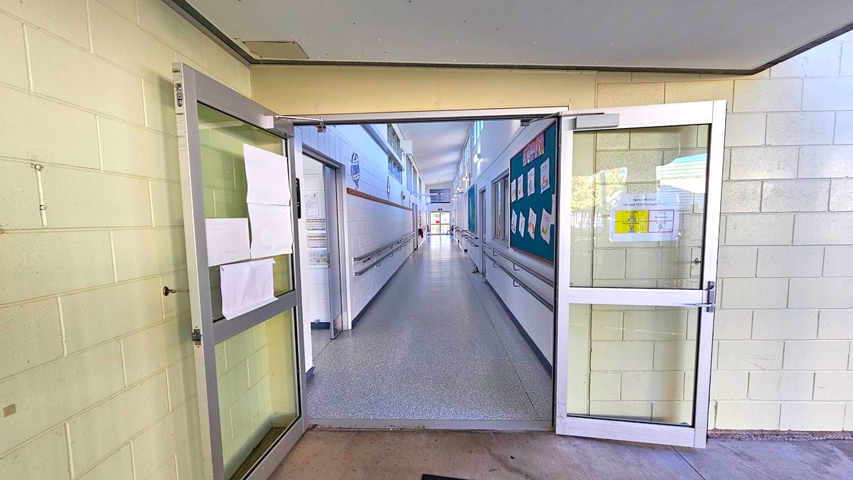 On the left, there is an open glass door with papers taped to it and a beige tiled wall. On the right, there is another open glass door with a green bulletin board displaying various notices and a beige tiled wall. In the middle, there is a long corridor with white walls, handrails, and a speckled grey floor, leading to a distant area. The ceiling is white with fluorescent lights. The floor at the entrance is concrete.