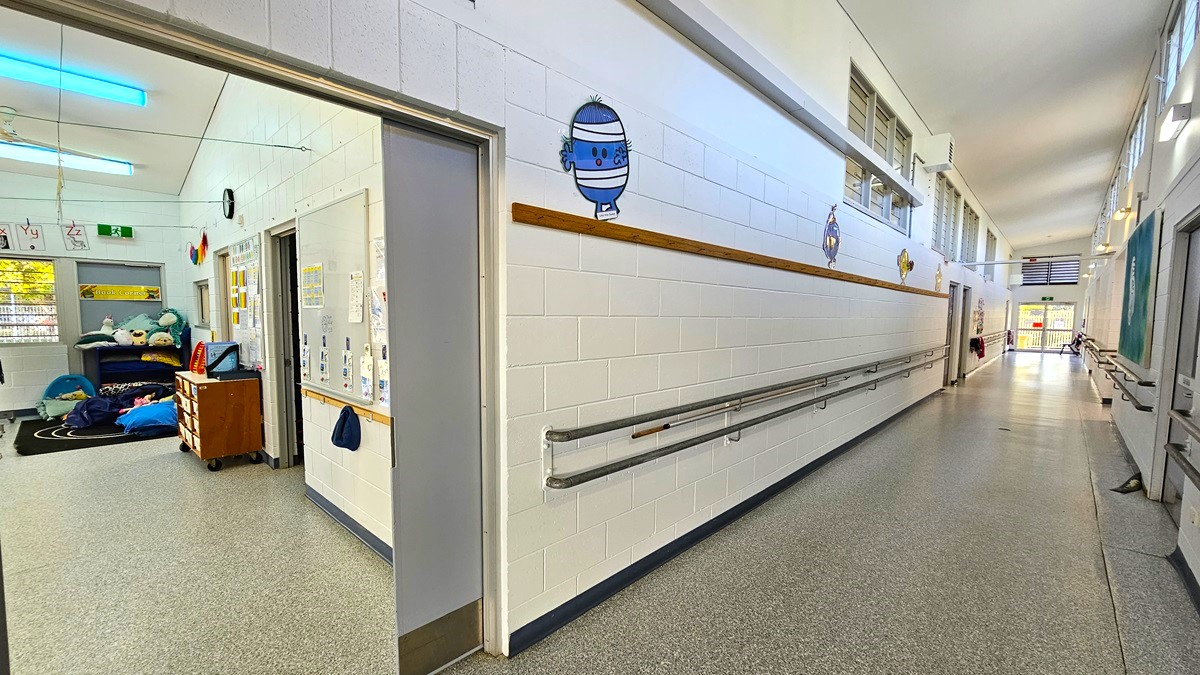 On the left, there is an open doorway leading to a room with a wooden dresser, blue cushions, and a black rug with toys. On the right, there is a long corridor with white walls, handrails, and windows near the ceiling. In the middle, the corridor extends into the distance with more windows and doors. The floor is speckled grey, and the ceiling is white with fluorescent lights and exposed beams.