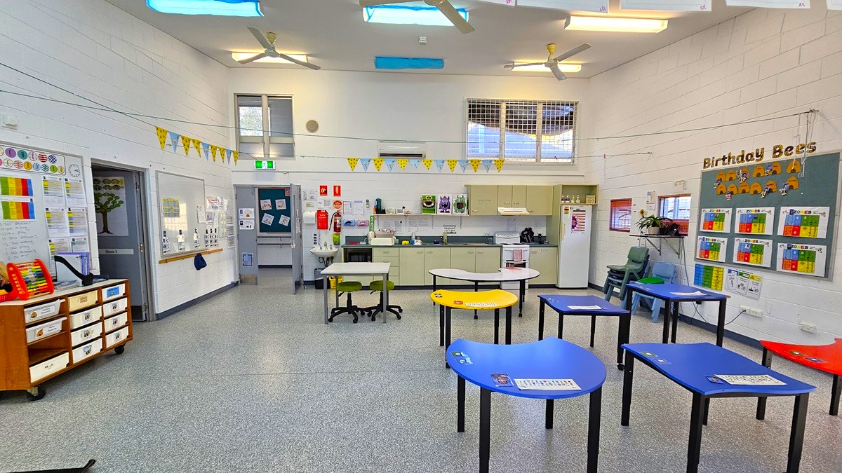 On the left, there is a wooden dresser with drawers, a whiteboard with colourful markers, and a door with posters. On the right, there is a bulletin board titled "Birthday Bees" with charts and a small sink area. In the middle, there are several blue and yellow tables with black legs, and a few chairs. The background features a kitchenette with cabinets, a sink, and a notice board. The floor is speckled grey, and the ceiling is white with skylights, fluorescent lights, and ceiling fans.