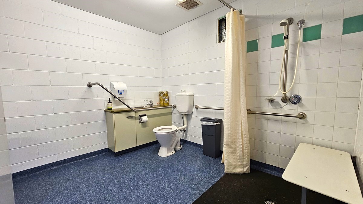 On the left, there is a white tiled wall with a grab bar, a paper towel dispenser, and a beige cabinet with a sink and cleaning supplies on top. On the right, there is a shower area with a white curtain, a showerhead, and grab bars. In the middle, there is a white toilet with a grey bin next to it. The floor is blue with a textured surface, and the ceiling is white with a vent and recessed lighting.