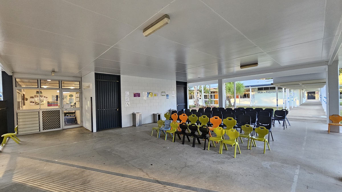 On the left, there is a glass door entrance with a metal screen door and a window displaying posters. In the middle, there are rows of colourful plastic chairs, mostly black and yellow, arranged in front of a white wall with a black door and a notice board. On the right, there is a long corridor with more chairs and a sign reading "Main Admin." The floor is concrete, and the ceiling is white with fluorescent lights. The background includes palm trees and additional buildings.