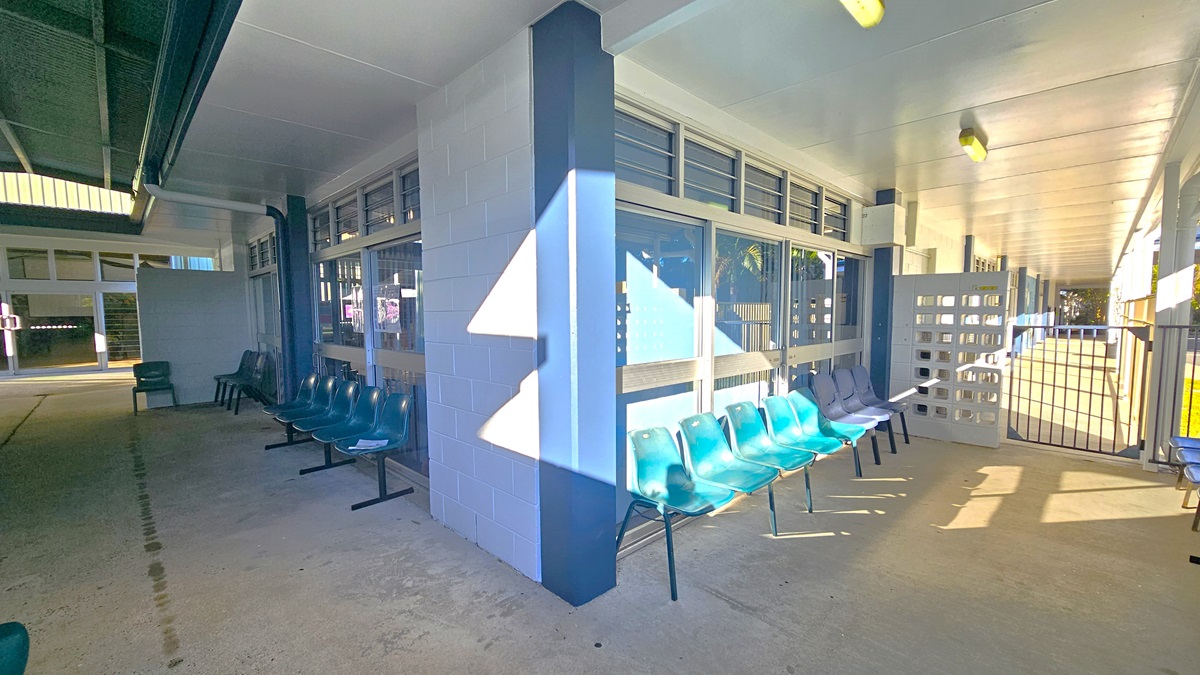 On the left, there are rows of black and green plastic chairs against a white wall with windows above. In the middle, there is a blue pillar with more green chairs arranged around it. On the right, there is a gated area with a white lattice fence and additional green chairs. The floor is concrete, and the ceiling is white with fluorescent lights. The background includes more windows and a corridor leading to other parts of the building.
