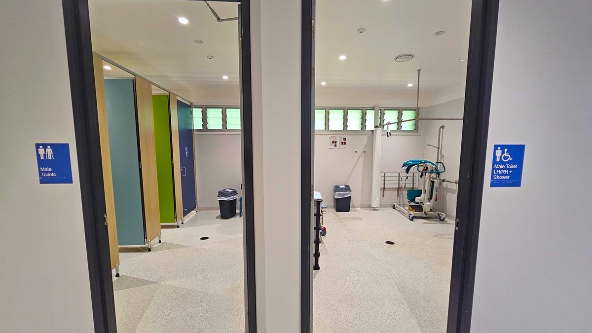 On the left, there is a doorway leading to a room with a blue sign indicating male toilets and showers. Inside, there are green and blue walls, a black bin, and a shower area. In the middle, there is a room with exercise equipment, a black bin, and a white wall with a few notices. On the right, there is another doorway with a blue sign indicating male toilets and showers, leading to a similar room with green and blue walls and a black bin.