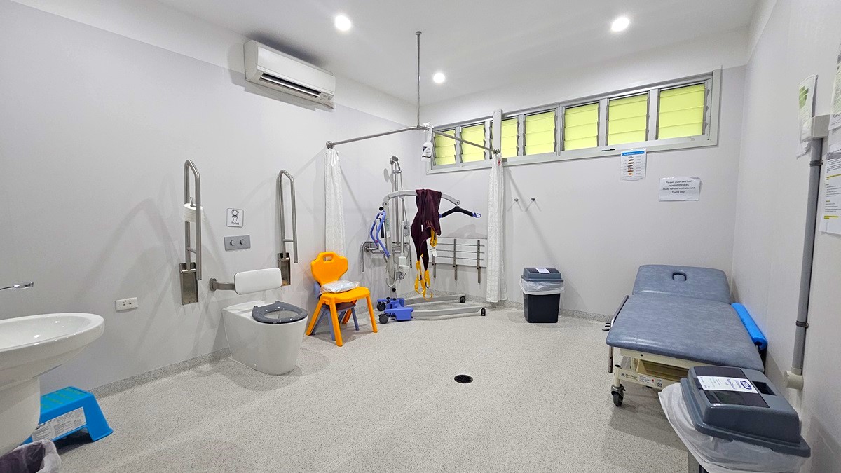 On the left, there is a white sink with a soap dispenser, a white toilet with support bars, and a blue box on the floor. In the middle, there is a patient lift machine with a harness, an orange chair, and a black bin. On the right, there is a grey examination table with a blue cushion, a small table with medical supplies, and a white wall with a few notices. The background has a row of frosted windows. The floor is light grey, and the ceiling has recessed lights and an air conditioning unit.
