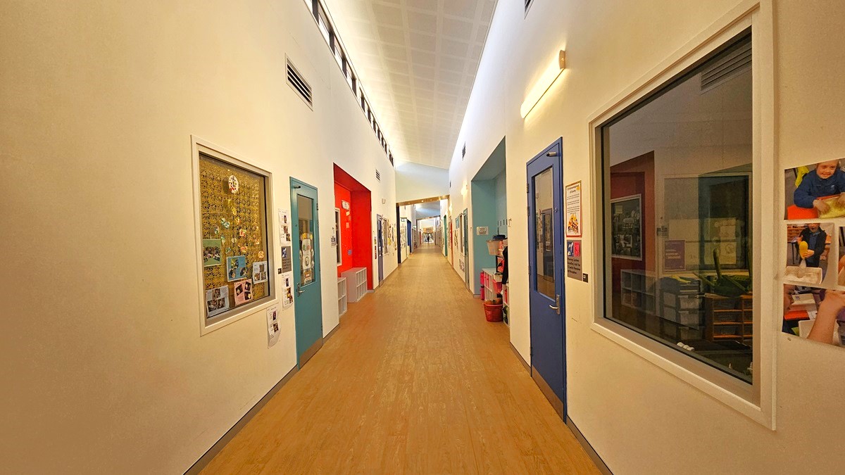 On the left, there are white walls with bulletin boards displaying various pictures and artwork. In the middle, there is a long hallway with light-coloured wooden flooring and a white, grid-patterned ceiling with recessed lighting. On the right, there are more bulletin boards, a blue door, and large windows showing rooms with furniture and decorations. The hallway extends into the distance with additional doors and windows on both sides.