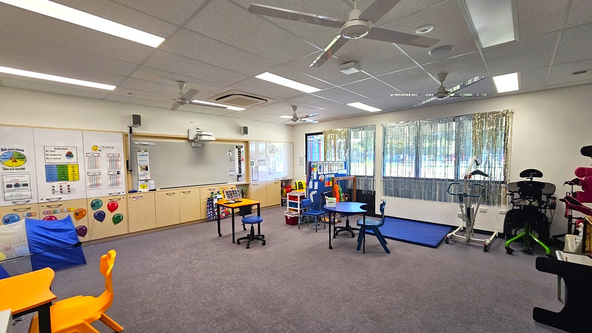 On the left, there are educational posters on the wall above a row of beige cabinets, a whiteboard, and a projector mounted on the ceiling. In the middle, there are small tables and chairs in blue and yellow, with a blue mat and a small bookshelf. On the right, there are large windows with vertical blinds, exercise equipment, and a blue exercise mat. The floor is grey carpet, and the ceiling is white with recessed lighting, ceiling fans, and an air conditioning unit.