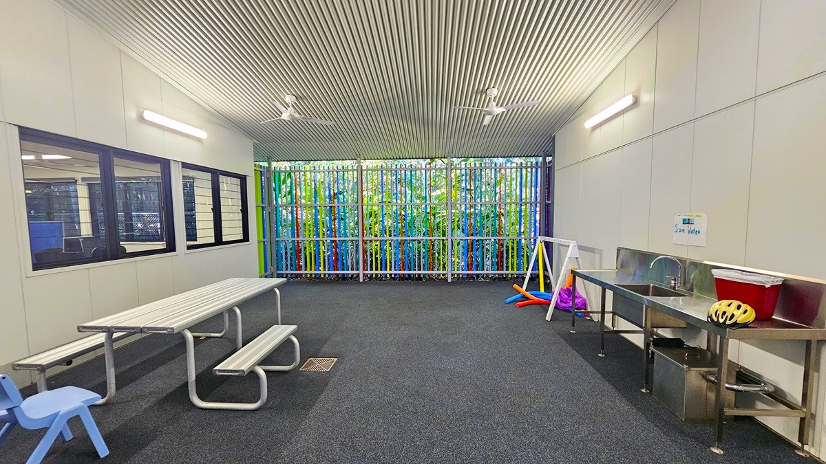 On the left, there are large windows with black frames, showing an adjacent room with tables and chairs. In the middle, there is a picnic table with attached benches and a small blue chair. On the right, there is a stainless steel sink with a countertop, a yellow helmet, and various colourful play items, including hula hoops and cones. The background features a wall of vertical, multicoloured bars. The floor is dark grey carpet, and the ceiling is white with linear grooves and fluorescent lights