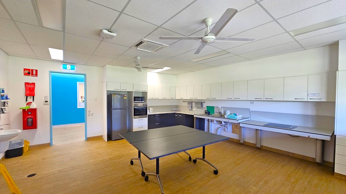 On the left, there is a white door with a blue exit sign above it, a fire extinguisher, and a fire hose reel mounted on the wall. In the middle, there is a black table with wheels on a wooden floor. On the right, there are white cabinets and countertops with a sink, microwave, and various items. The ceiling has white tiles, fluorescent lights, ceiling fans, and an air conditioning vent.