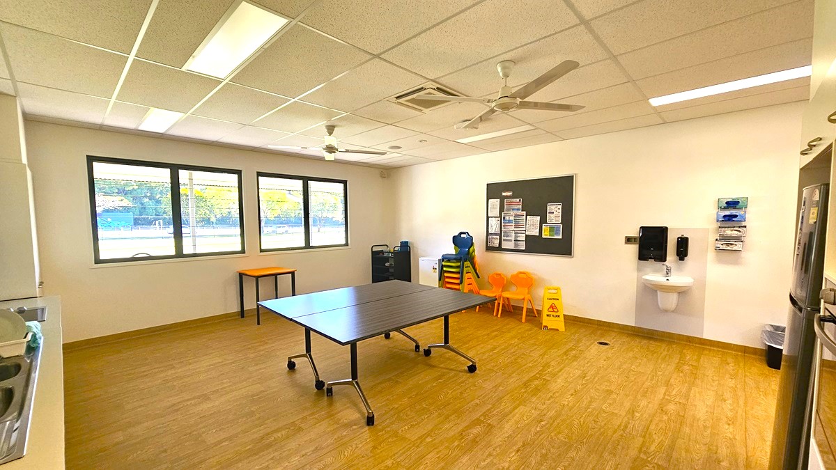 On the left, there are three large windows with black frames. In the middle, there is a black table with wheels on a wooden floor. On the right, there is a wall-mounted sink with soap dispensers and a hand dryer above it, a bulletin board with various notices, and a row of orange chairs next to a small orange table. The ceiling has white tiles, fluorescent lights, ceiling fans, and an air conditioning vent.
