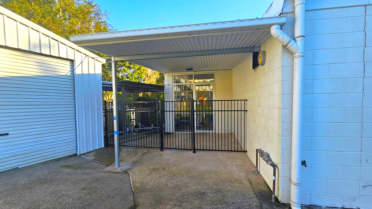 On the left, there is a white building with a metal roof and several windows, surrounded by a black fence. In the middle, there is a grassy area with a concrete slab and a tall light pole. On the right, there is a white building with a blue door and a small window, with trees in the background. The foreground is a large green lawn.