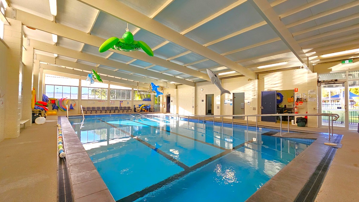 On the left, there are colourful inflatable toys and a row of chairs against the wall. In the middle, there is an indoor swimming pool with lane markers and floating inflatable animals, including a green turtle and a blue dolphin. On the right, there are more chairs, a black door, and a fire extinguisher mounted on the wall. The floor is tiled around the pool, and the ceiling has exposed beams with fluorescent lights and skylights. The background includes large windows letting in natural light.