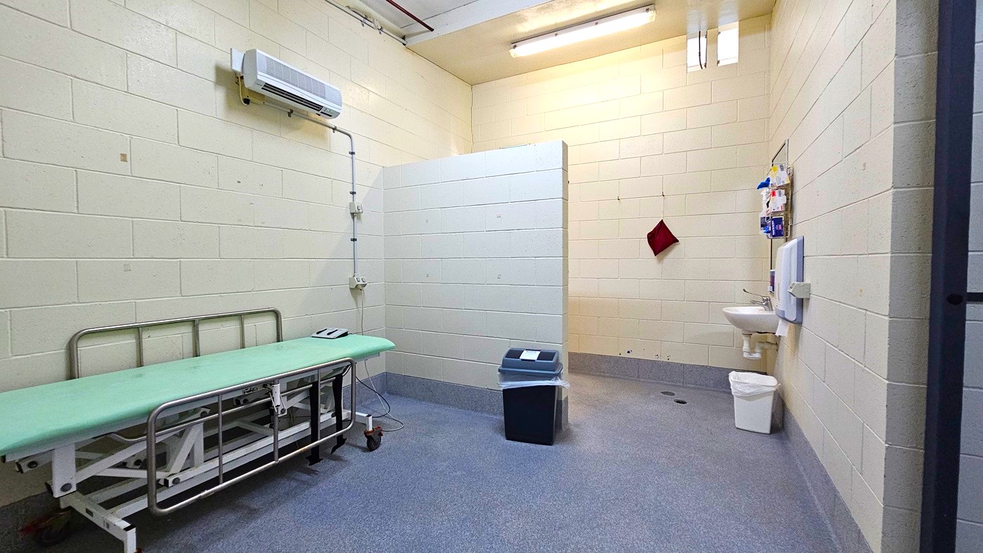 On the left, there is a green cushioned examination table on wheels with a control panel, and an air conditioning unit mounted on the wall above it. In the middle, there is a partial wall divider and a blue bin on the floor. On the right, there is a white sink with a mirror, a soap dispenser, a red towel hanging on a hook, and a white bin below. The floor is grey with a drainage hole, and the ceiling has exposed pipes and fluorescent lights. The background includes cream-coloured brick walls.