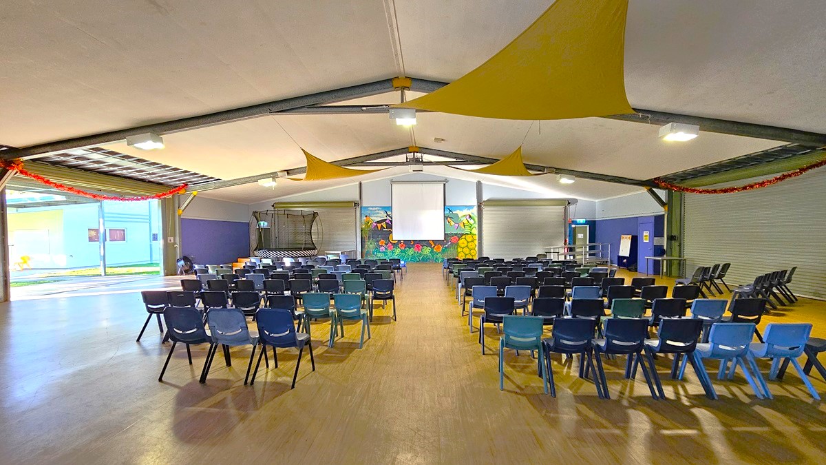On the left, there is a trampoline with a safety net and a small stage area with a blue door. In the middle, there are rows of black and blue chairs facing a projector screen and a colourful mural of a garden on the back wall. On the right, there are two large roller doors and a blue wall with a few chairs stacked against it. The floor is wooden, and the ceiling is white with exposed metal beams and yellow fabric draped across, along with fluorescent light fixtures.