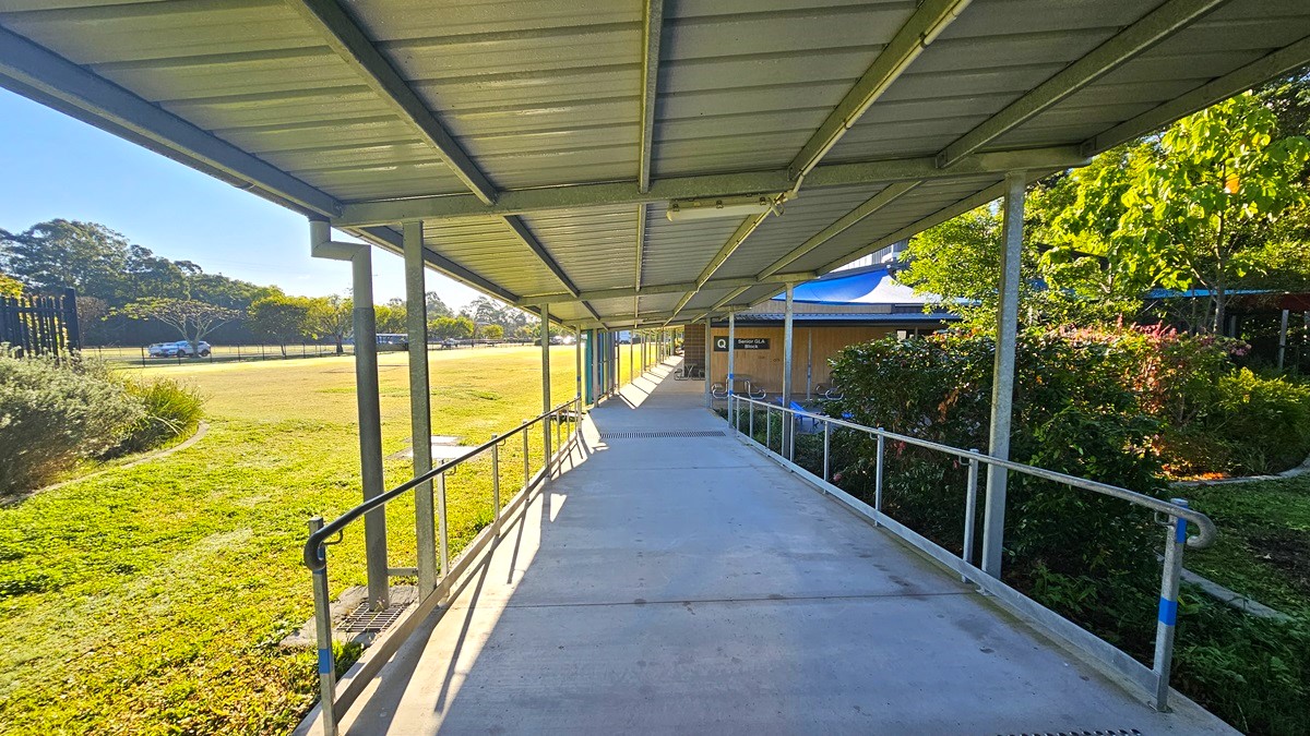 On the left, there is a grassy field with trees and a fence in the distance. In the middle, there is a long concrete walkway with metal railings, covered by a corrugated metal roof supported by grey poles. On the right, there are bushes and a building with a blue roof partially visible. The floor is concrete, and the ceiling is a corrugated metal roof.
