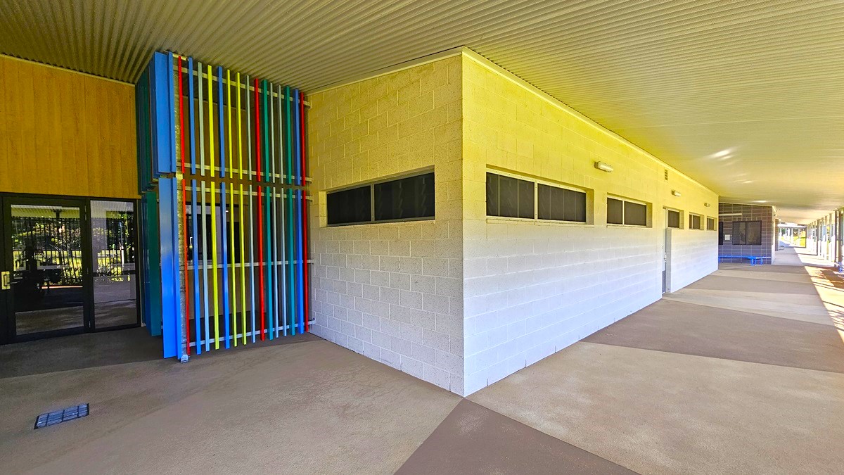 On the left, there is a building entrance with glass doors and a colourful structure made of vertical blue, yellow, red, and green bars. In the middle, there is a long beige wall with small rectangular windows near the top. On the right, the wall continues with more windows and a blue door in the distance. The floor is a mix of beige and brown concrete, and the ceiling is a white corrugated metal roof.