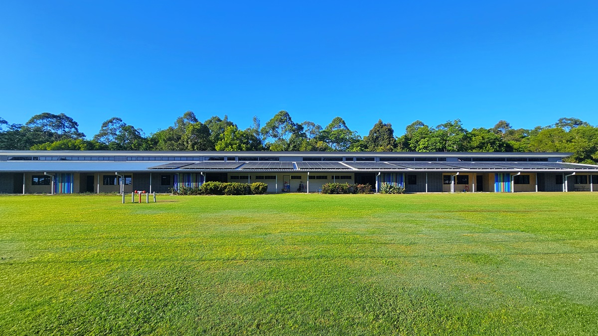 On the foreground, there is a grassy field with a few small wooden structures. In the middle, there is a long, single-storey building with a dark roof, featuring multiple windows and doors, and a row of bushes in front. On the right, the building continues with more windows and doors, and a blue door is visible. The floor is a well-maintained grassy field, and the ceiling is a dark, flat roof. In the background, there are tall trees behind the building.