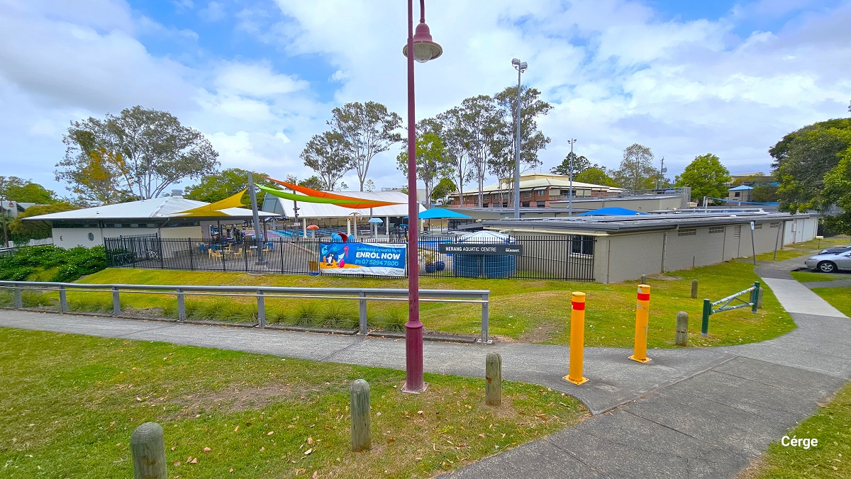 Arrival at Nerang Aquatic Centre from Martin Street with yellow curb marking along an intersection. The sloping road has a middle red brick section and is connected to a middle red and white road markings. Opposite the road is the facility with a small number of designated car spaces marked by white lines, which are occupied by varying brands and colors and cars.