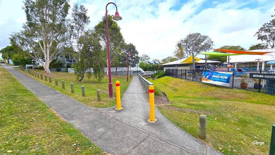 View standing on the start of the concrete pathway with two yellow bollards leading to the entrance and reception desk of Nerang Aquatic Centre. The right side is lined with grey metal handrails with strings of wire underneath, while the left side is red bell-shaped lamp posts. The left section is mostly occupied by grass and tall trees. Hanging on the black perimeter fence is a large blue advertisement and a green signboard detailing the name of the centre.