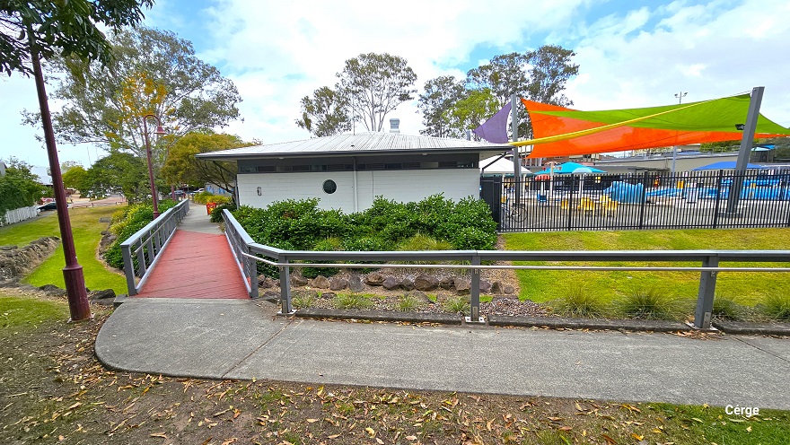 Image shows a red bridge section with grey guardrails connected from the concrete pathway. On both sides is a beautiful landscape of well-maintained grass and a shrub above an arranged rock. Right after the red bridge section is the administration office of a small crème brick building with a prominent hole in the wall. Visible from this position is the colorful sun shades covering the pool.