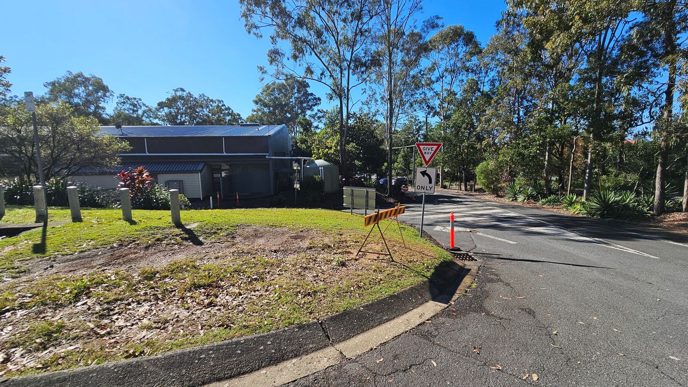 A road curves to the right with a "Give Way" sign and a directional arrow indicating a left turn only. On the left, a building with a metal roof is partially visible, surrounded by trees and shrubs. Concrete bollards line the grassy area in front of the building. A temporary barrier and an orange traffic cone are placed near the road. Tall trees with sparse foliage are in the background, and the road surface is slightly cracked with scattered leaves.