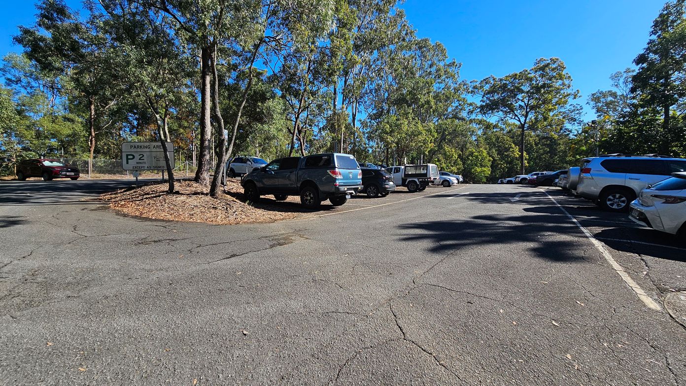 A car park surrounded by tall trees. On the left, a sign indicates a 2-minute parking area. Several vehicles, including a blue SUV and a white utility vehicle, are parked on the left side. On the right, more cars are lined up along the edge of the asphalt surface. The ground is cracked and uneven, with scattered leaves. The background is filled with dense greenery, and the sky is clear and blue.