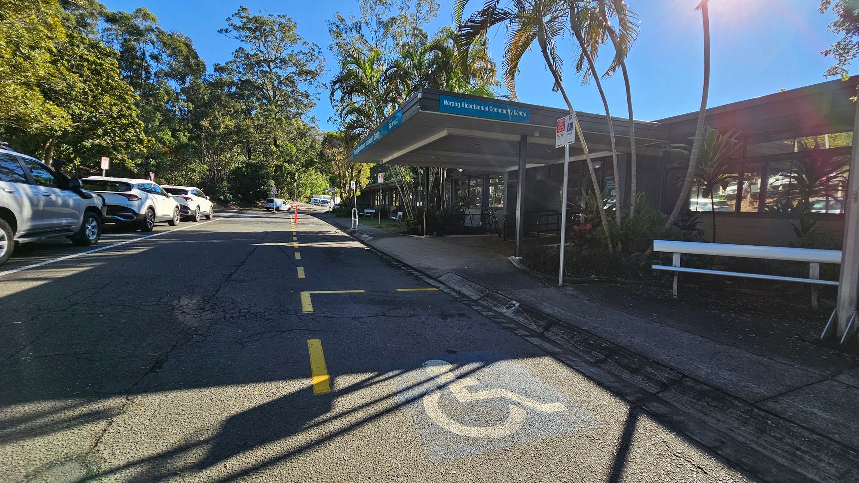A street scene with a row of parked cars on the left side, including several white vehicles. On the right, a building with a sign reading "Nerang Bicentennial Community Centre" is partially visible, with a covered entrance and a bench nearby. Palm trees and other greenery surround the area. The road has a marked disabled parking space in the foreground, and yellow lines are painted on the asphalt. The background features more trees and a clear blue sky.