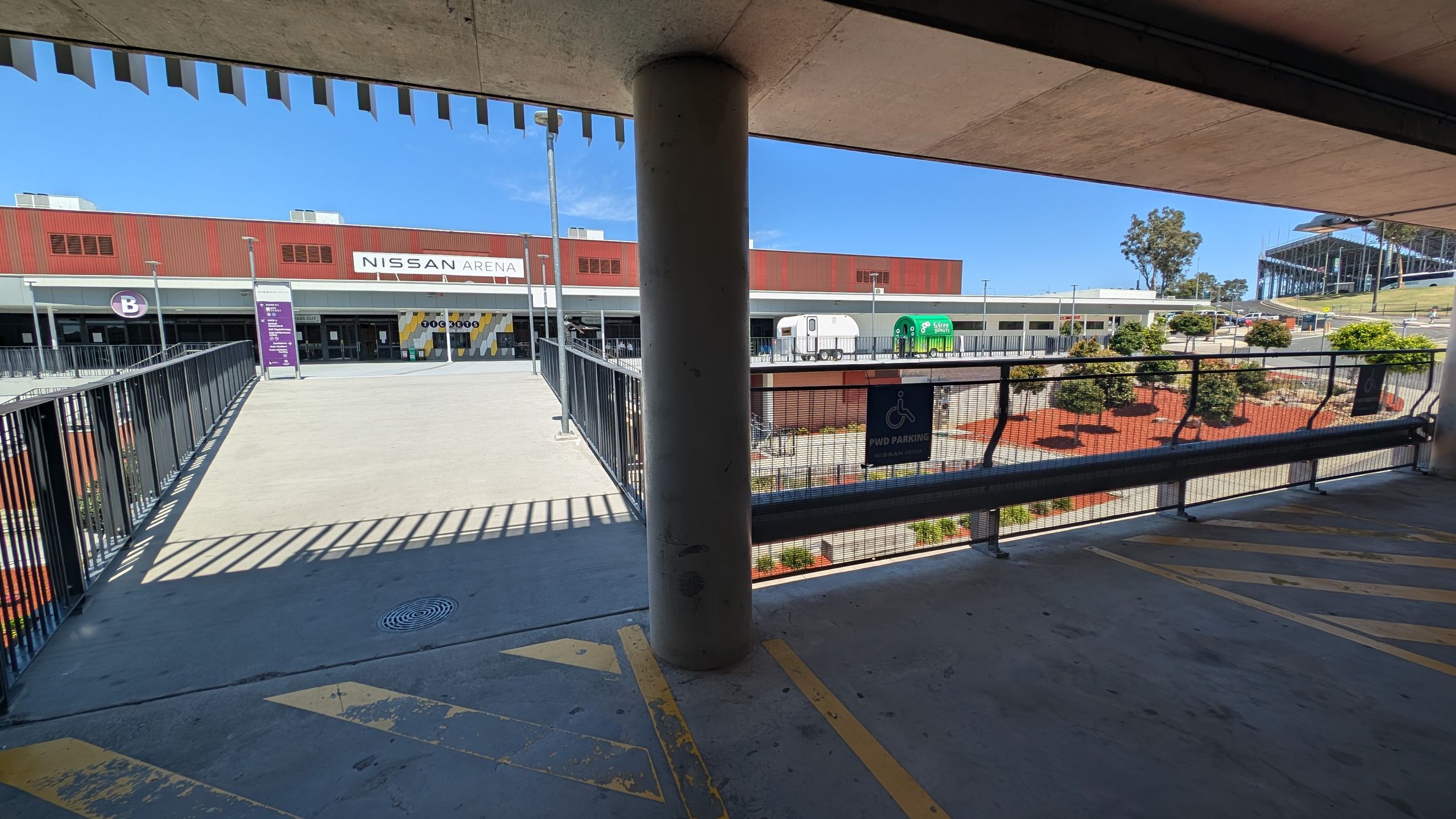 A wide view of the Nissan Arena entrance. The building has a red and white facade with a large sign reading "Nissan Arena" at the top centre. Below, there are entrances marked with letters "B" and "C". A statue of a person holding a ball is on the right side in the foreground. The ground is paved, and there are some outdoor seating areas with umbrellas near the entrances.