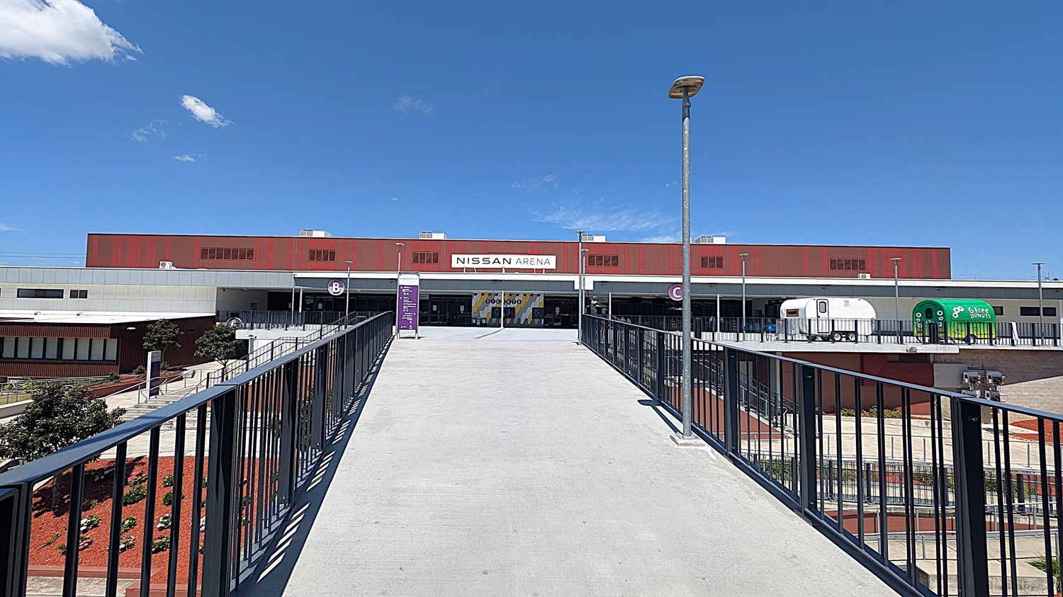 A wide concrete walkway leads to the entrance of a building with a sign reading "Nissan Arena". The building has a red and white facade. On the right, there are two trailers, one white and one green with "free donuts" written on it. The walkway is flanked by black railings and lit by street lamps. On the left, there are landscaped areas with small plants and trees.