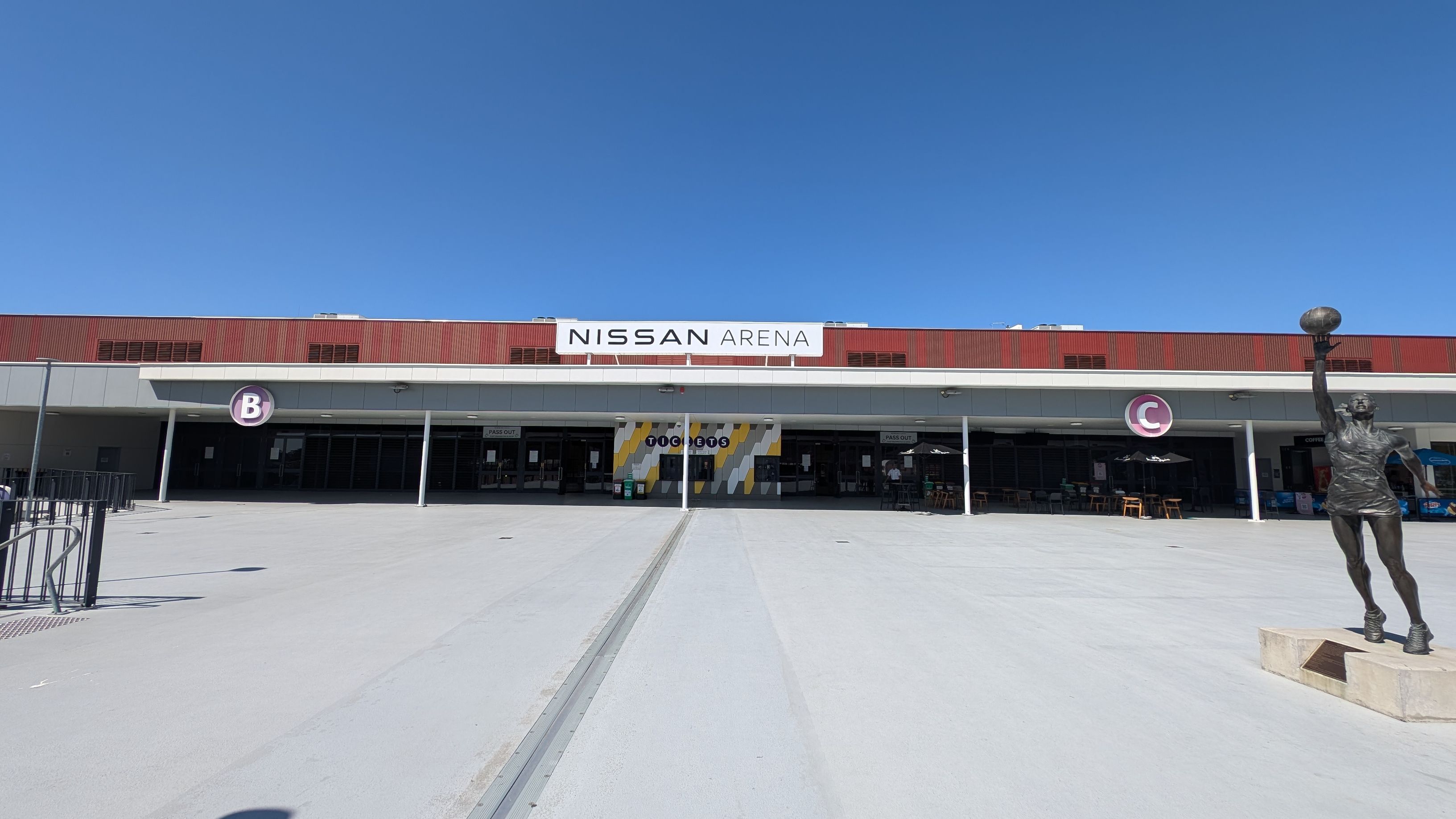A large building with a sign reading "Nissan Arena" on the right. In front, there are several parking spaces marked with blue disabled signs and yellow lines. A white and red barrier gate is visible in the foreground. On the left, a covered walkway with white pillars extends towards the background. There are a few trees along the building's side, and the sky is clear with a few clouds.