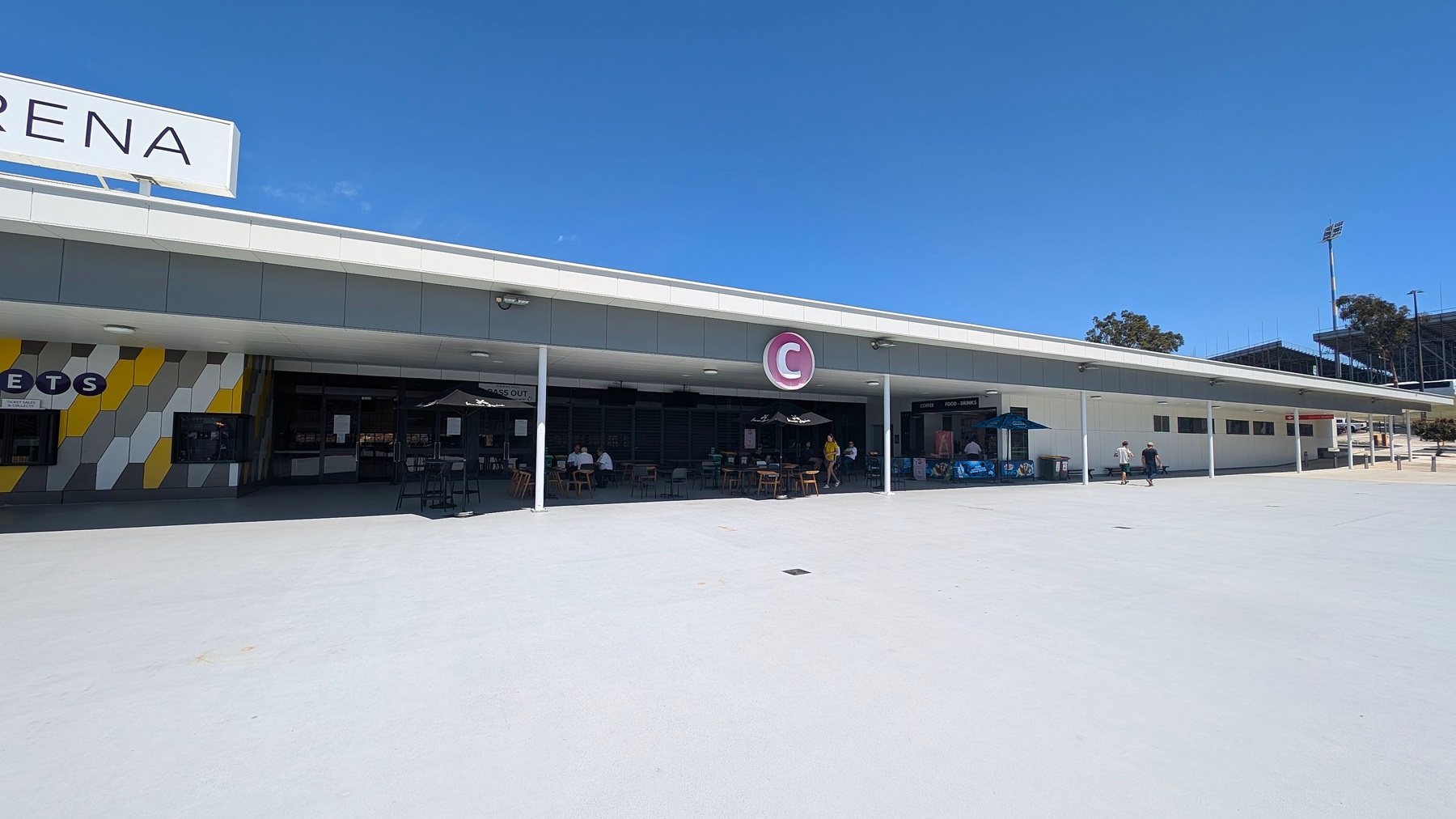 A wide, open area with a modern building in the background. The building has a flat roof and a large letter "C" sign. On the left, there is a section with a geometric pattern in grey, yellow, and white, labelled "TICKETS". Several tables with umbrellas are set up outside, with a few people seated. To the right, there are more people walking near a food and beverage stand. The ground is a smooth, light-coloured surface.