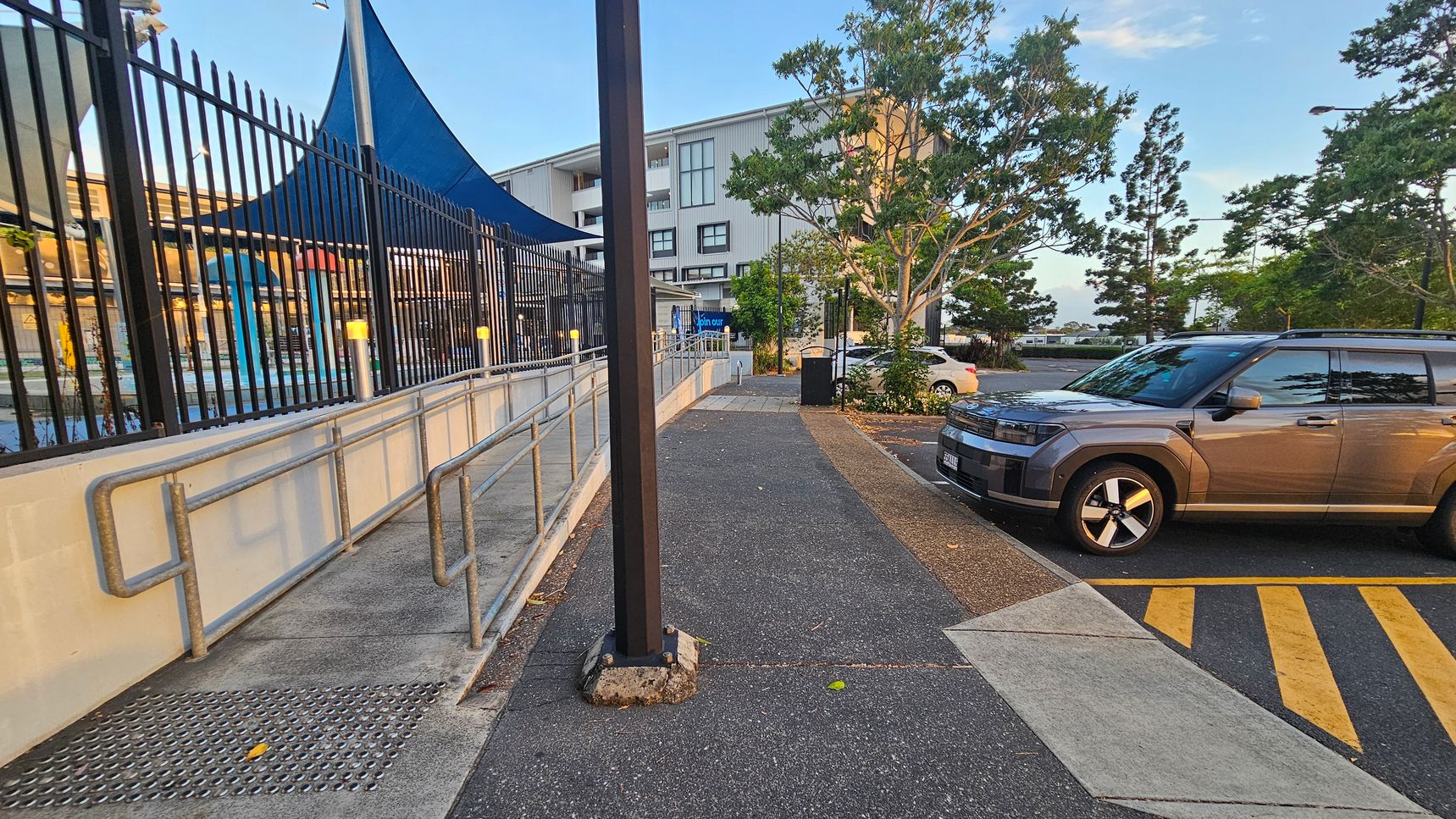 A pathway runs through the centre, bordered by a metal fence on the left with a blue shade structure above, likely covering a pool area. A metal handrail follows the path. On the right, a grey car is parked in a marked bay with yellow lines. Trees and a multi-storey building are visible in the background. A tall pole stands in the middle of the path. The ground is a mix of concrete and asphalt.