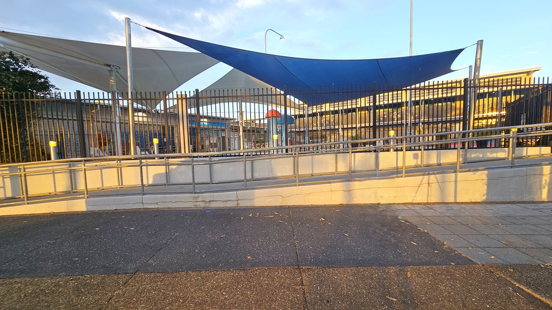 A paved area with a mix of dark and light grey tiles in the foreground. A metal railing and a concrete ramp lead up to a fenced area. Behind the fence, there are large triangular shade sails in blue and white, supported by metal poles. A building with a corrugated metal roof is visible in the background, along with a colourful play structure. The scene is lit by warm sunlight, casting long shadows.