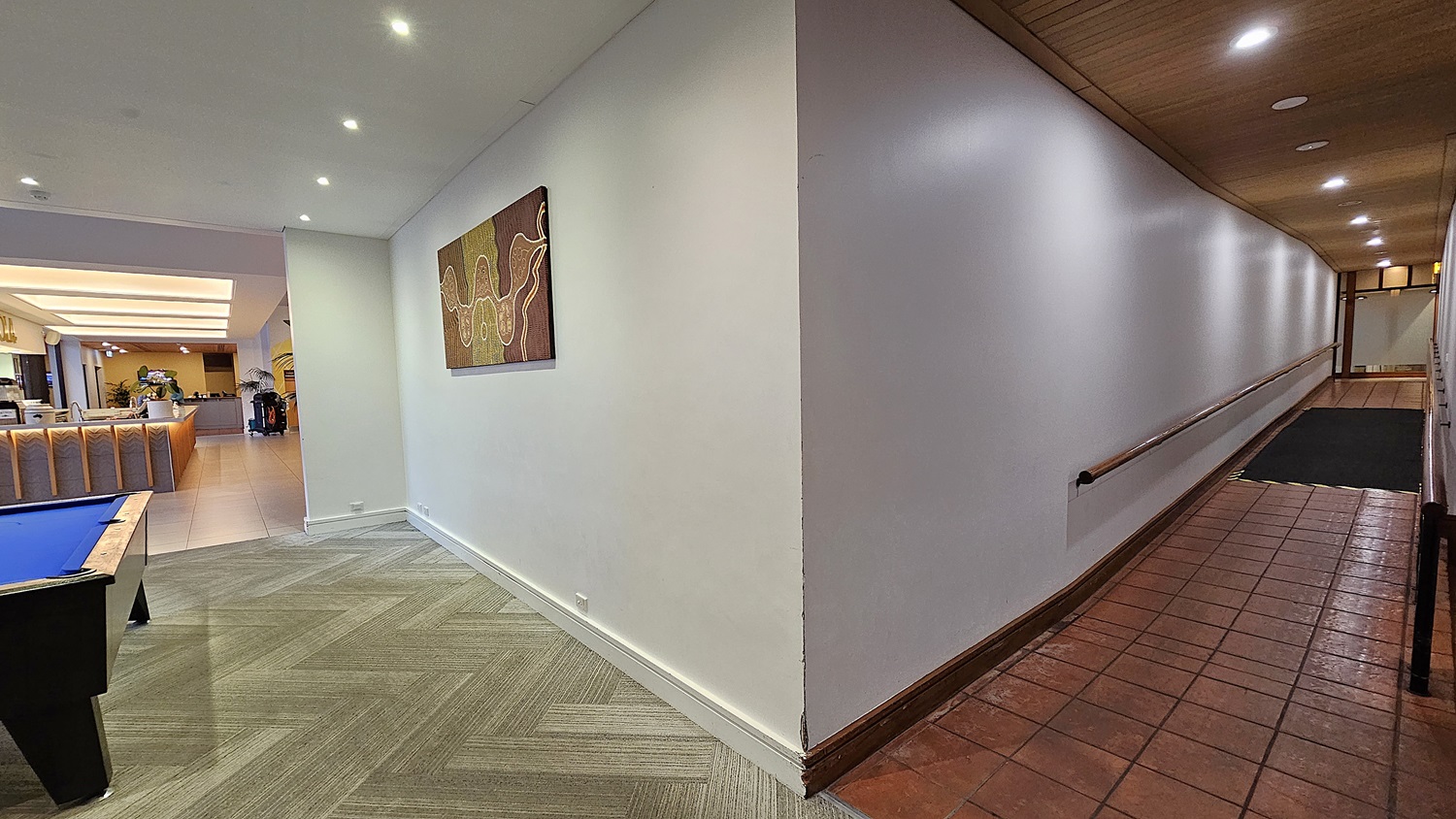 On the left, there is a pool table with a blue surface next to a white wall adorned with a colourful abstract painting. To the right, a long hallway with terracotta floor tiles and a wooden ceiling features a handrail along the white wall. In the background, a well-lit area with a reception desk and seating arrangements is visible. The floor transitions from patterned carpet to light tiles, and the ceiling has recessed lighting, creating a bright and open space.
