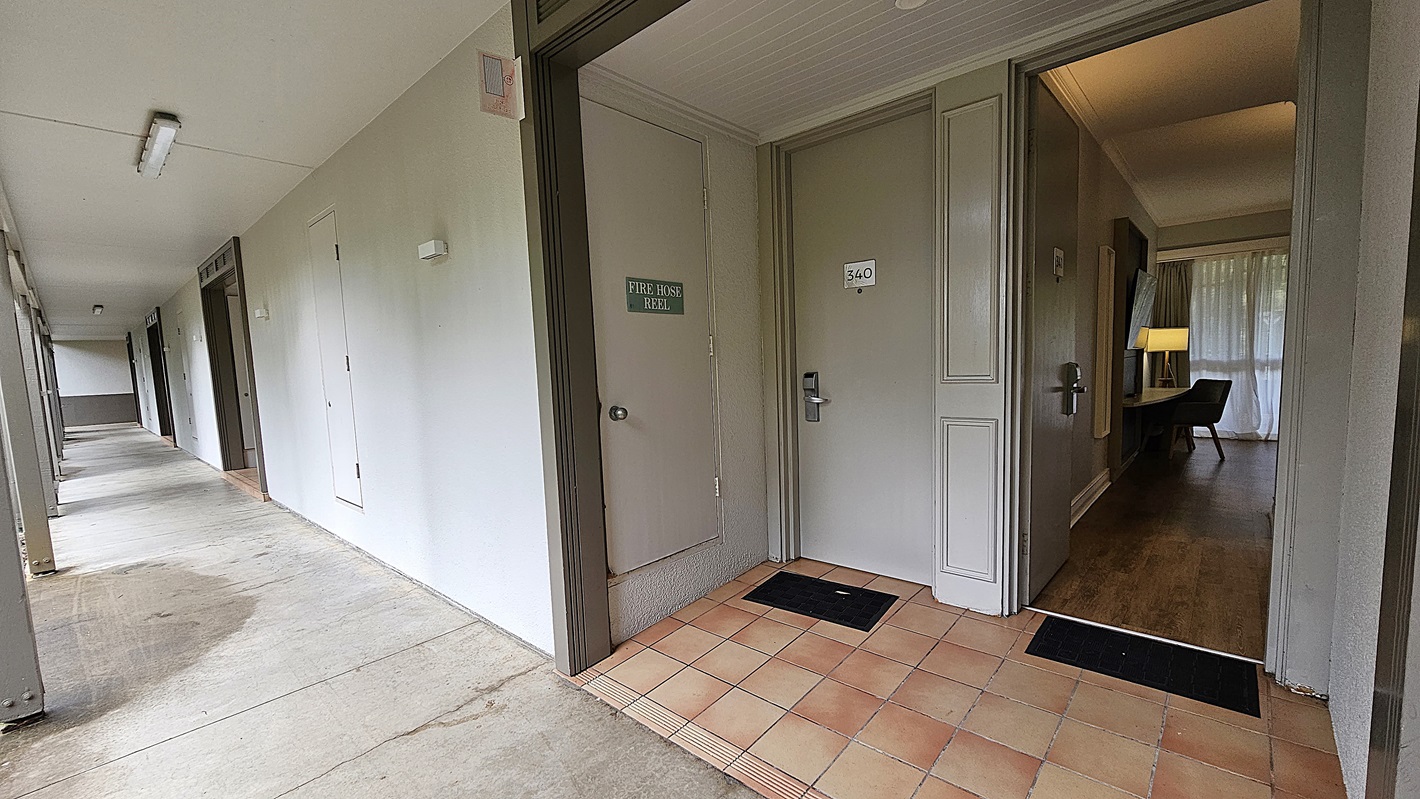 On the left, a long corridor with white walls and grey doors extends into the distance, with a fire hose reel mounted on the wall. To the right, an open door reveals a room with wooden flooring, a desk, a chair, and a lamp. In the middle, the corridor and the entrance to the room meet at a tiled area with a black doormat. The floor is a mix of concrete and tiles, and the ceiling is flat and white with fluorescent lights.