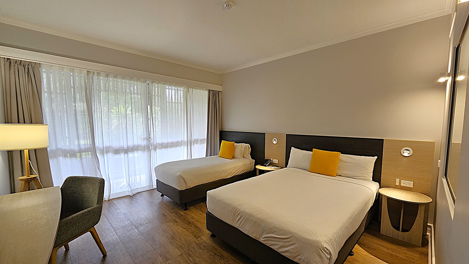 On the left, a large window with sheer curtains and grey drapes, next to a wooden table with a lamp and a grey upholstered chair. To the right, two beds with white linens and yellow cushions, each with a wooden bedside table and wall-mounted reading lights. In the middle, the beds are positioned against a grey wall with a dark wooden headboard. The floor is wooden, and the ceiling is white with recessed lighting.