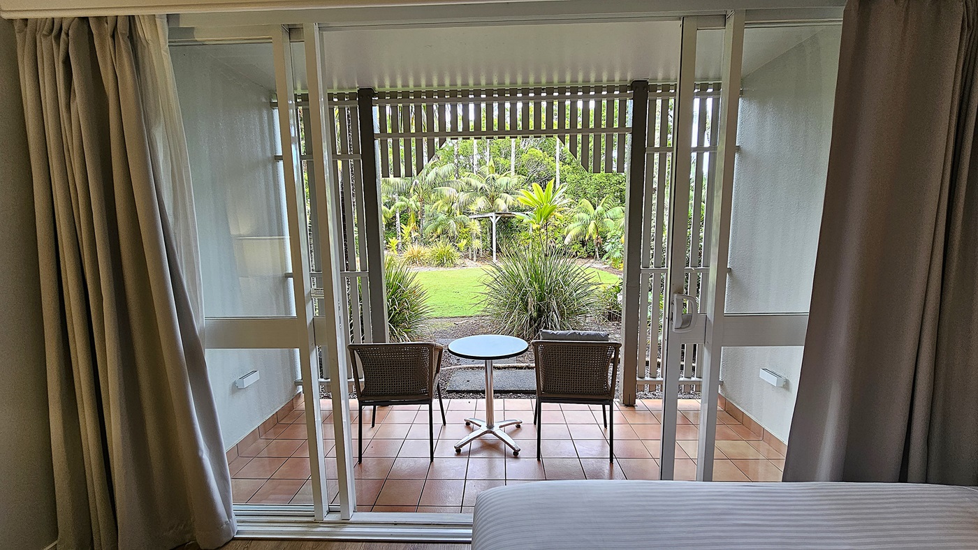 On the left, beige curtains partially drawn open. To the right, more beige curtains. In the middle, a glass sliding door opens to a patio with two wicker chairs and a round table. Beyond the patio, a garden with green plants and palm trees. The floor inside is tiled, and the ceiling is white.