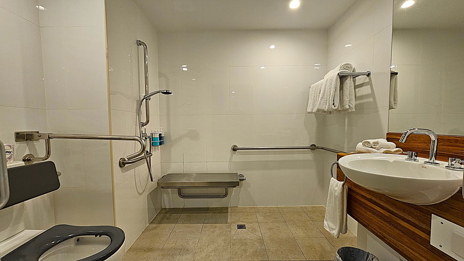 On the left, a toilet with a black seat and a wall-mounted grab bar. To the right, a white sink with a chrome faucet set in a wooden countertop, with folded white towels on a rack above. In the middle, a wall-mounted shower seat and a handheld showerhead with soap dispensers. The floor is tiled in beige, and the ceiling is white with recessed lighting.