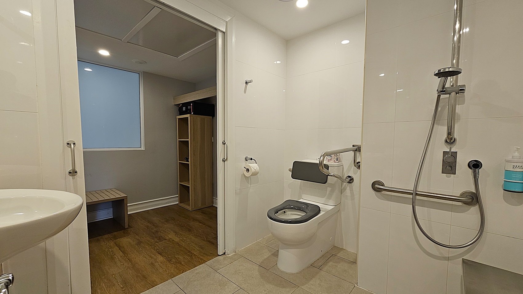 On the left, a white sink with a chrome faucet. To the right, a toilet with a grey seat, a wall-mounted grab bar, and a handheld showerhead with a soap dispenser. In the middle, an open doorway leads to a room with wooden flooring, a wooden bench, and a shelving unit. The floor in the bathroom is tiled in beige, and the ceiling is white with recessed lighting.