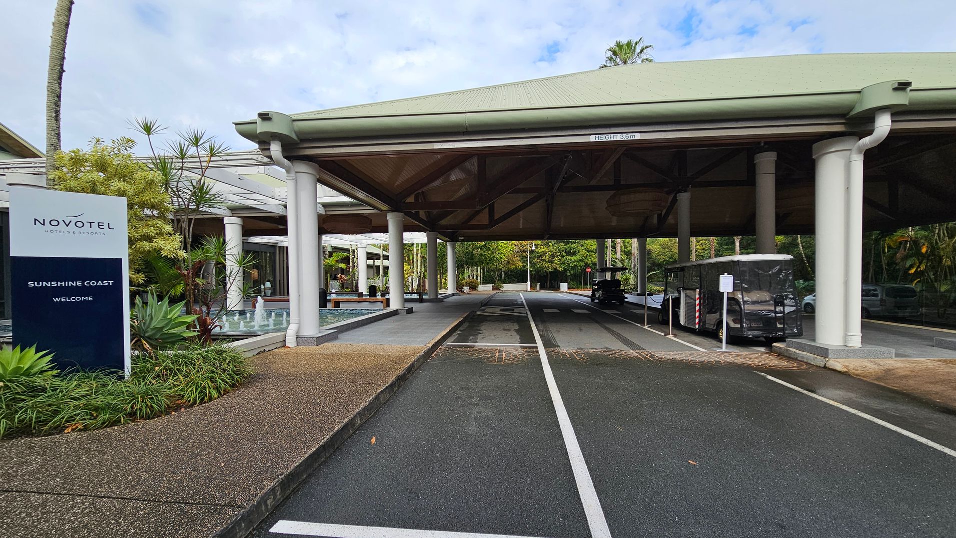 A covered driveway with a green roof supported by white pillars. On the left, there's a sign for Novotel Hotels & Resorts, Sunshine Coast, surrounded by lush greenery and a small fountain. The driveway has marked lanes, and a covered vehicle is parked on the right. The background features more greenery and a pathway leading further into the area. The ground is a mix of asphalt and textured paving.