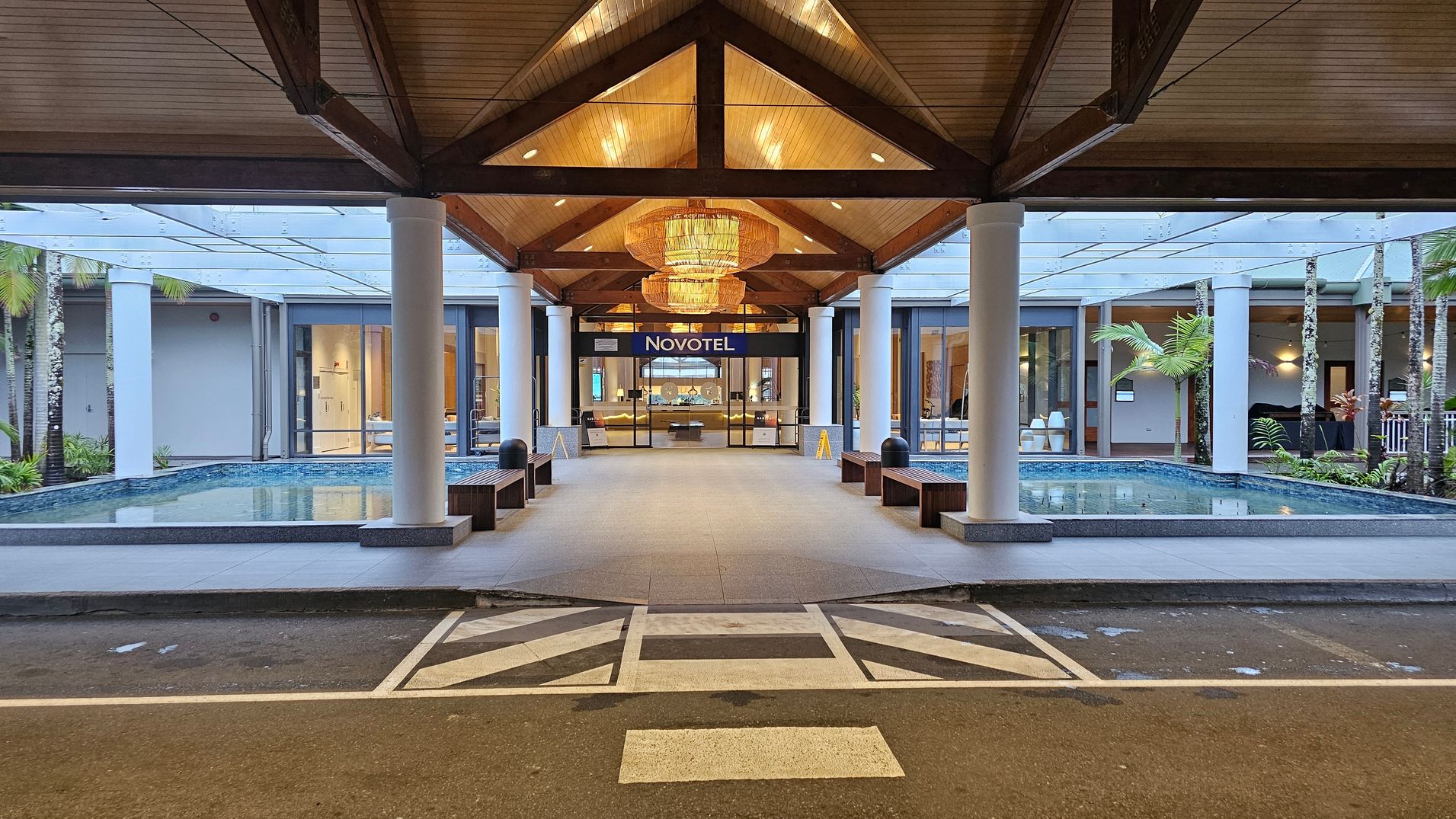 A hotel entrance with a covered walkway leading to glass doors marked "Novotel". The ceiling is wooden with large chandeliers. On either side of the walkway are shallow pools of water, bordered by white columns. Benches are placed along the sides. The background shows a modern interior with glass walls and visible seating areas. The foreground has a marked pedestrian crossing on the road.