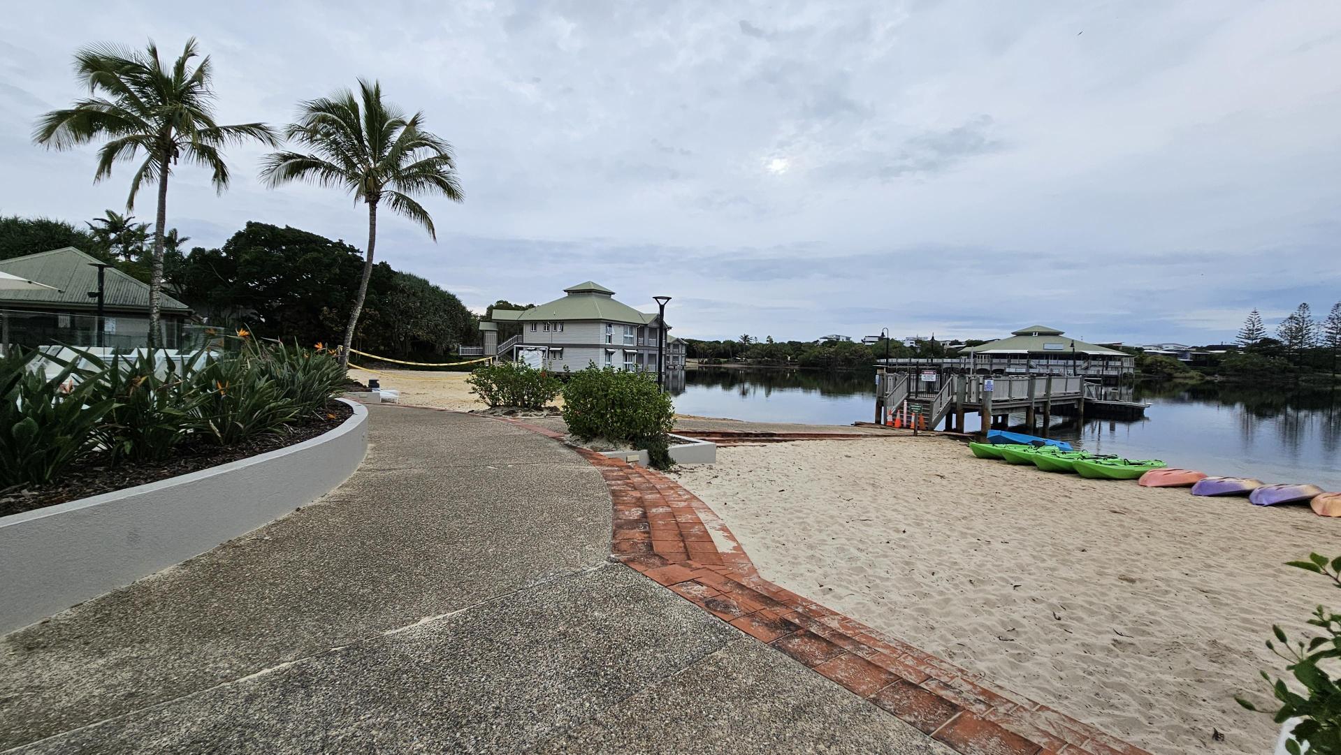 Sandy beach in the foreground with a volleyball net set up. On the left, a building with a green roof is partially visible. In the centre, a calm body of water reflects the surrounding structures and trees. A wooden pier extends into the water. On the right, a tall palm tree stands next to a building with a similar green roof. The ground near the building is paved with a mix of green and grey stones, bordered by red bricks.