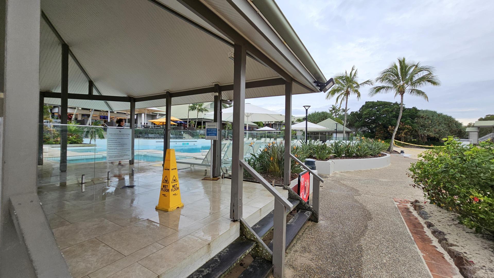 A poolside area with a wet floor sign on a tiled surface under a covered structure. Glass barriers surround the pool, and there are steps leading down to a textured path. On the right, there are palm trees and lush greenery. In the background, there are more covered seating areas with umbrellas and additional greenery.